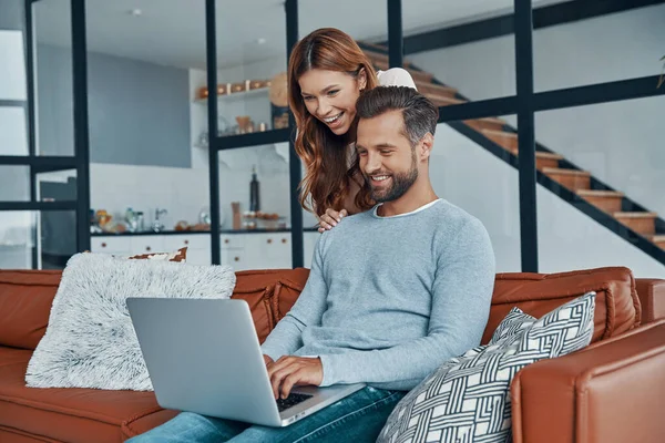 Jovem casal alegre usando laptop e sorrindo enquanto passa o tempo em casa — Fotografia de Stock