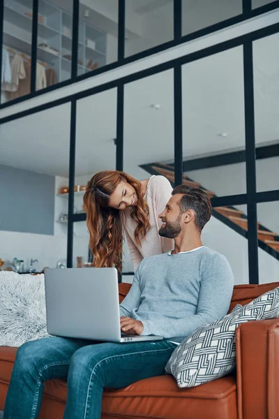 Amar jovem casal usando laptop e sorrindo enquanto passa o tempo em casa — Fotografia de Stock