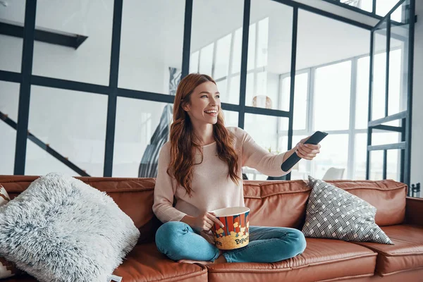 Красивая молодая женщина смотрит телевизор и ест попкорн, сидя — стоковое фото