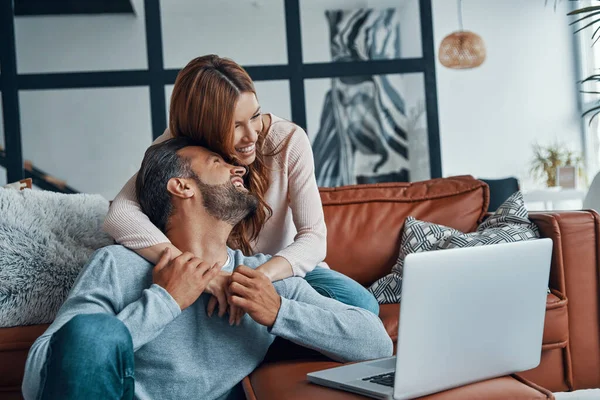 Belo jovem casal usando laptop e sorrindo enquanto passa o tempo em casa — Fotografia de Stock