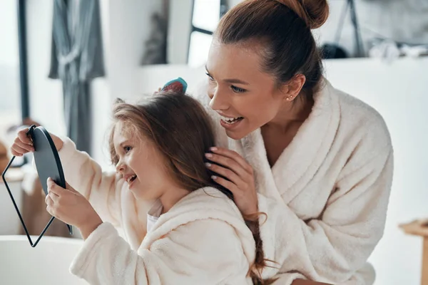Schöne junge Mutter kombiniert ihre Tochter Haare, während sie Morgenroutine macht — Stockfoto