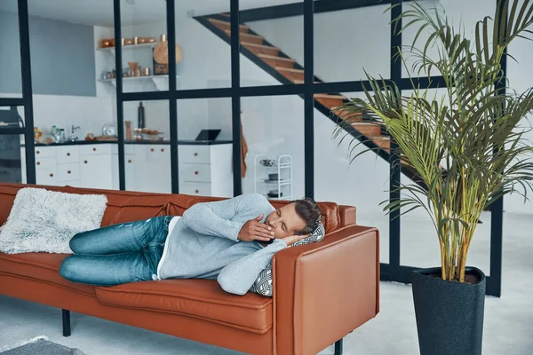 Przystojny młody człowiek w luźnych ubraniach ziewa leżąc na kanapie w domu — Zdjęcie stockowe