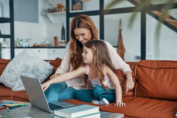 Moeder en haar dochter hechting samen en glimlachen tijdens het gebruik van laptop thuis — Stockfoto