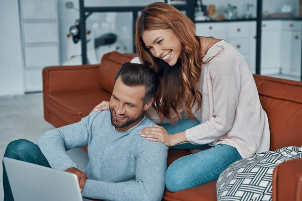 Ευτυχισμένο νεαρό ζευγάρι που χρησιμοποιεί φορητό υπολογιστή και χαμογελά, ενώ περνούν το χρόνο τους στο σπίτι — Φωτογραφία Αρχείου