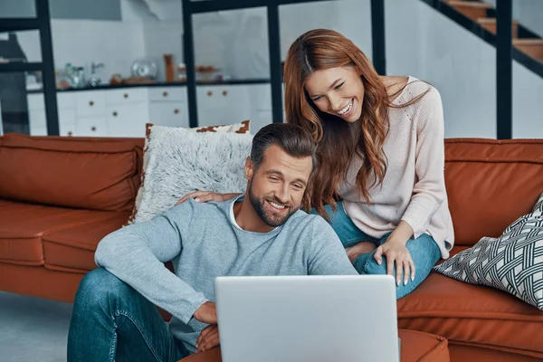 Belo jovem casal usando laptop e sorrindo enquanto passa o tempo em casa — Fotografia de Stock