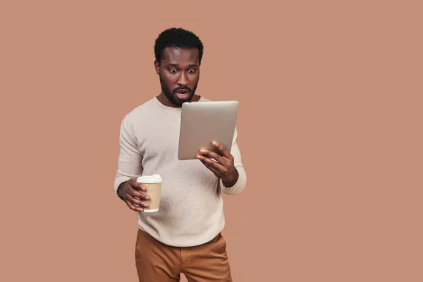 Bonito jovem africano em roupas casuais usando tablet digital e segurando xícara de café — Fotografia de Stock