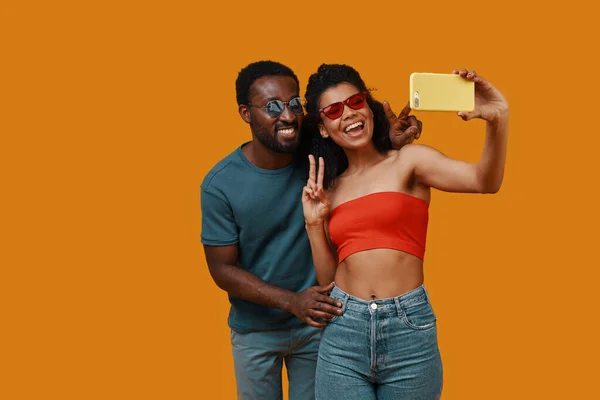 Hermosa joven pareja africana sonriendo y tomando selfie mientras está de pie sobre el fondo amarillo — Foto de Stock