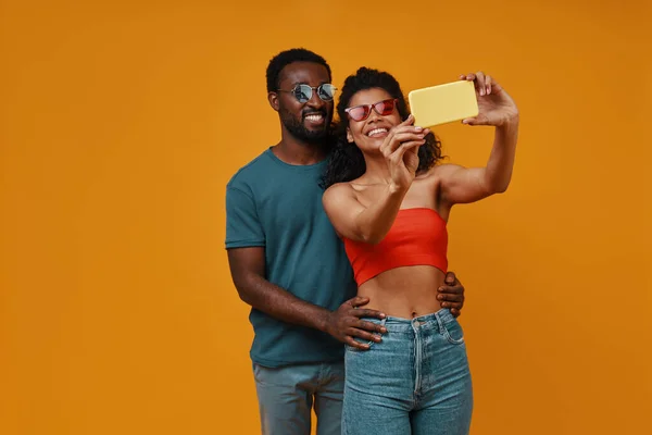 Hermosa joven pareja africana sonriendo y tomando selfie mientras está de pie sobre el fondo amarillo — Foto de Stock