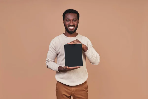 Przystojny młody Afrykanin w luźnej odzieży pokazujący przestrzeń do kopiowania na tablecie cyfrowym i uśmiechnięty — Zdjęcie stockowe