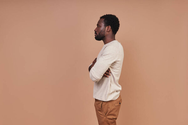 Side view of handsome young African man in casual holding arms crossed while standing against brown background