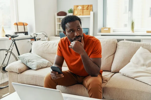 Pensativo joven africano que utiliza el teléfono inteligente y mantener la mano en la barbilla mientras pasa tiempo en casa — Foto de Stock