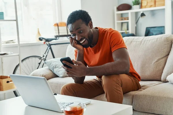 Bonito jovem africano usando telefone inteligente e sorrindo enquanto passa o tempo em casa — Fotografia de Stock