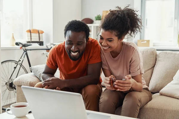 Piękna młoda afrykańska para patrząca na laptopa i uśmiechnięta podczas spędzania czasu w domu — Zdjęcie stockowe