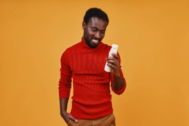 Yakışıklı Afrikalı genç adam, sarı arka planda dururken şişeye bakıyor ve gülümsüyor.