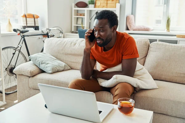 Jóképű afrikai férfi beszél telefonon és mosolyog, miközben otthon tölti az idejét. — Stock Fotó