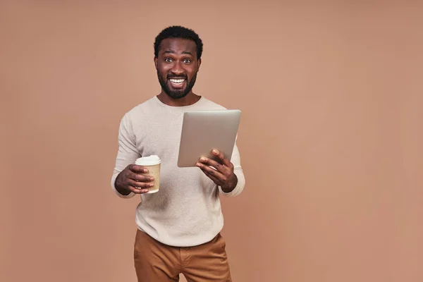 Joven hombre africano alegre en ropa casual sosteniendo tableta digital y taza de café — Foto de Stock