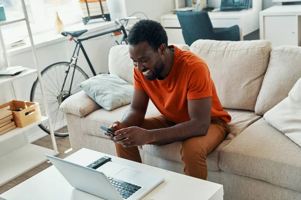 Joyeux jeune homme africain utilisant un téléphone intelligent et souriant tout en passant du temps à la maison — Photo