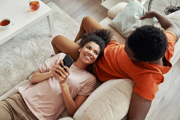 Vista dall'alto della giovane donna africana che usa lo smartphone e sorride mentre trascorre del tempo a casa con il suo ragazzo — Foto Stock