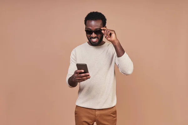 Stilig ung afrikansk man i avslappnade kläder med hjälp av smart telefon och leende — Stockfoto