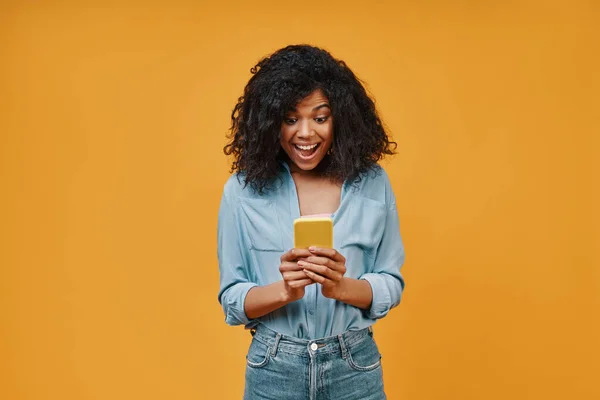 Hermosa mujer africana joven en ropa casual usando teléfono inteligente y sonriendo —  Fotos de Stock