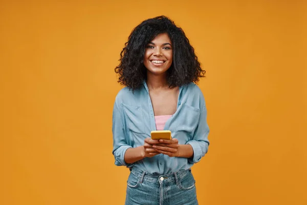 Güzel, genç Afrikalı bir kadın, günlük kıyafetler içinde, akıllı telefon kullanıyor ve gülümsüyor. — Stok fotoğraf
