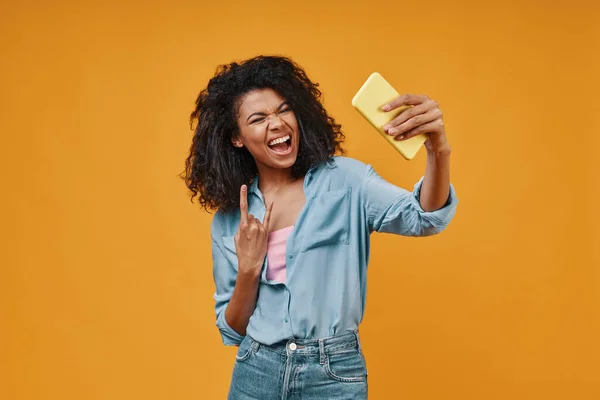 Playful young African woman in casual clothing taking selfie and gesturing while standing against yellow background — Stock Photo, Image
