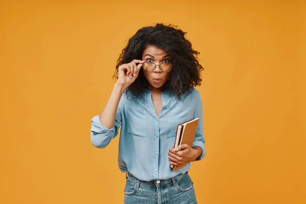 Mujer africana joven conmocionada llevando almohadillas de notas y ajustando las gafas mientras está de pie sobre el fondo amarillo — Foto de Stock