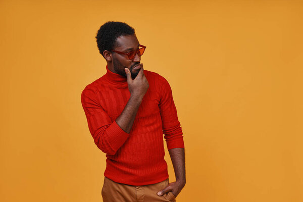 Stylish young African man in eyeglasses looking away while standing against yellow background.