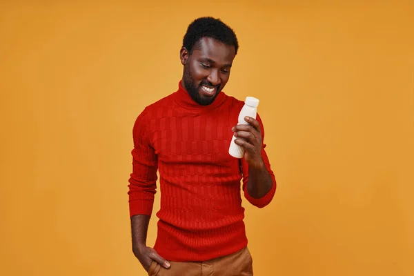 Knappe jonge Afrikaanse man die naar de fles kijkt en lacht terwijl hij tegen een gele achtergrond staat — Stockfoto