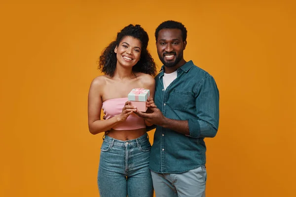 Όμορφο νεαρό ζευγάρι Αφρικής κρατώντας κουτί δώρου και χαμογελώντας, ενώ στέκεται πάνω σε κίτρινο φόντο — Φωτογραφία Αρχείου