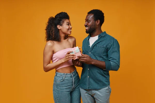 Красивые молодые африканские мужчины дарят подарочную коробку своей девушке, стоя на жёлтом фоне — стоковое фото