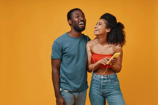 Hermosa joven pareja africana sonriendo y escuchando música mientras está de pie sobre el fondo amarillo — Foto de Stock