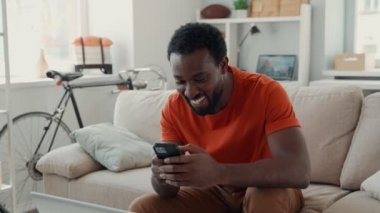 Genç Afrikalı adam akıllı telefon kullanıyor ve evde vakit geçirirken gülümsüyor.