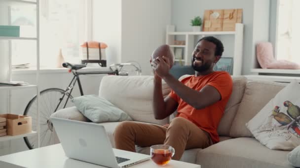 Feliz joven africano animando y sosteniendo la pelota de fútbol mientras ve el deporte — Vídeo de stock