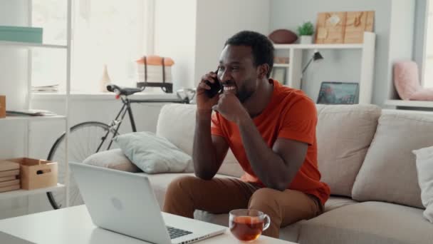 Молодой африканский мужчина разговаривает по телефону и улыбается, проводя время дома — стоковое видео
