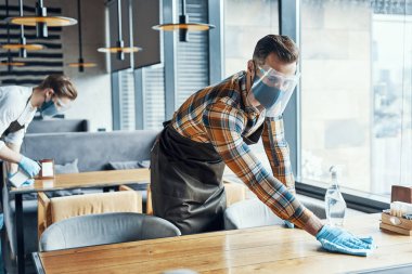 Two young male waiters in protective workwear cleaning tables in restaurant
