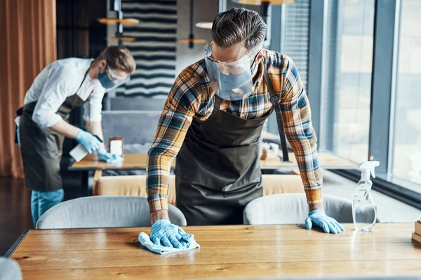Two male waiters in protective workwear cleaning tables in restaurant — Stockfoto