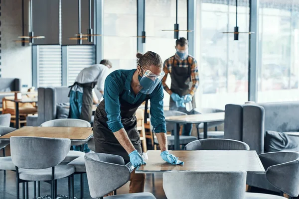 Tre unga manliga servitörer i skyddsarbetskläder rengöringsbord för kunder i restaurang — Stockfoto