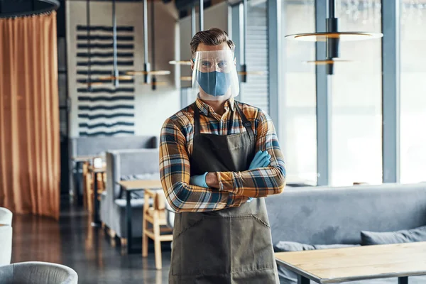 Male waiter in protective workwear keeping arms crossed and looking at camera while standing in restaurant - Stock-foto