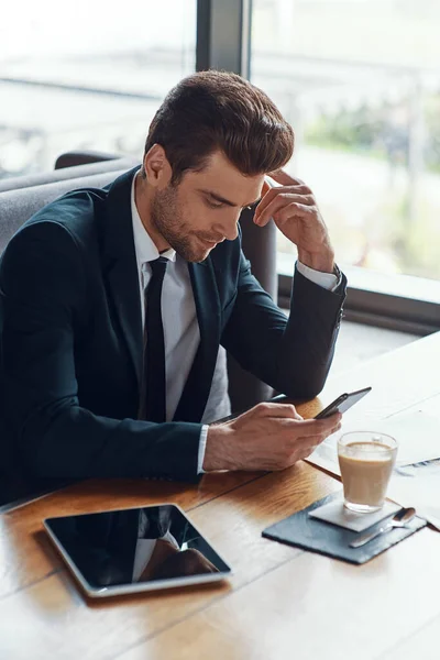Busy young businessman in full suit using smart phone while sitting in the restaurant — Stock Photo, Image