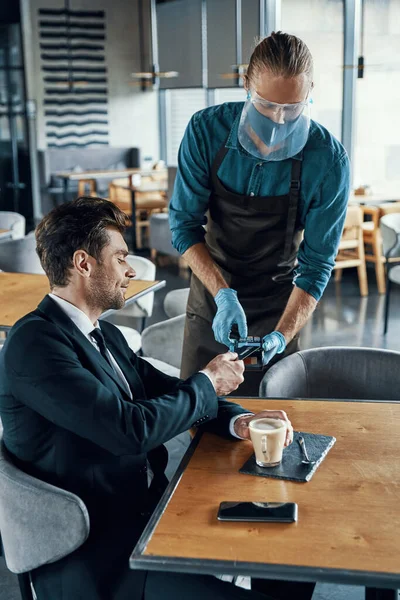 Bonito jovem empresário fazendo um pagamento sem contato enquanto sentado no restaurante — Fotografia de Stock