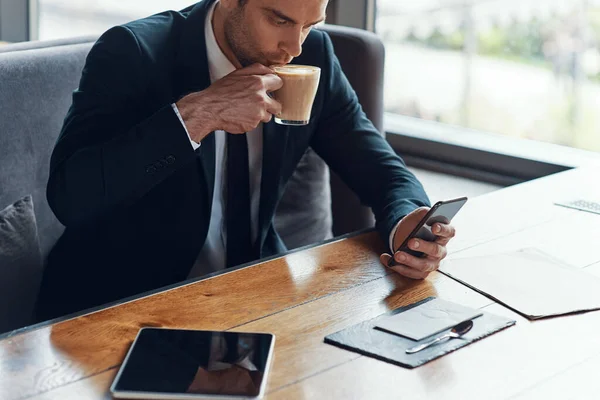 Handsome young businessman in full suit using smart phone and drinking coffee while sitting in the restaurant — Stock Photo, Image