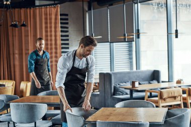 Handsome young men in aprons arranging furniture while preparing restaurant to opening clipart