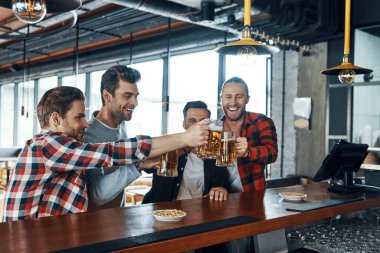 Happy young men in casual clothing toasting each other with beer and smiling while sitting in the pub