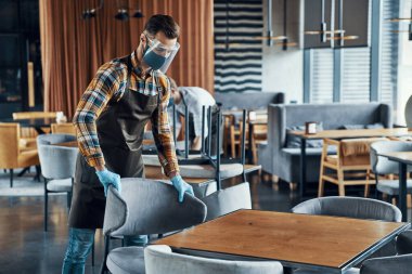 Young men in protective face shields and aprons arranging furniture in restaurant