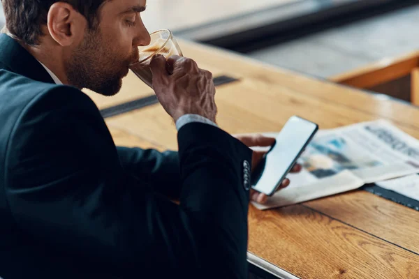 Handsome young businessman in full suit using smart phone and drinking coffee while sitting in the restaurant — Stock fotografie