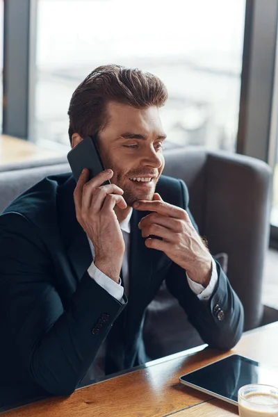 Sonriente joven hombre de negocios en traje completo hablando en el teléfono inteligente mientras está sentado en el restaurante — Foto de Stock