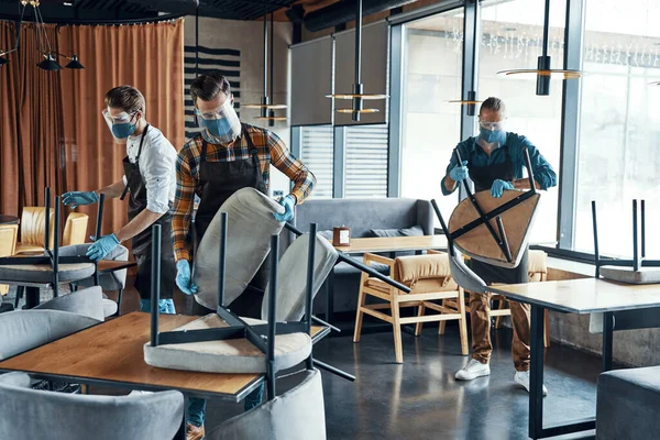 Три чоловічі офіціанти в захисному робочому одязі розставляють меблі в ресторані — стокове фото