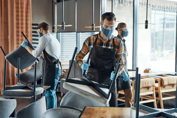 Busy young male waiters in protective workwear arranging furniture in restaurant — Stock Photo, Image