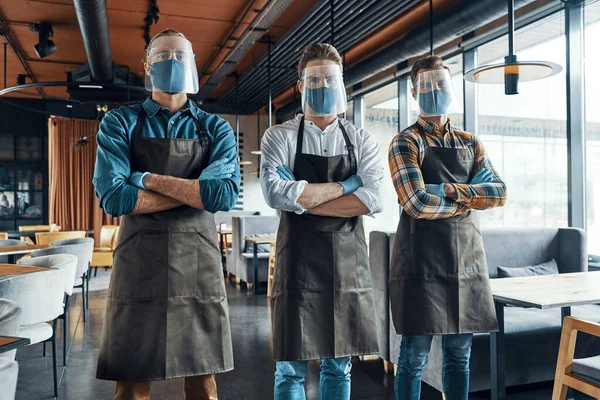 Three confident male waiters in protective workwear keeping arms crossed and looking at camera — Stock Photo, Image